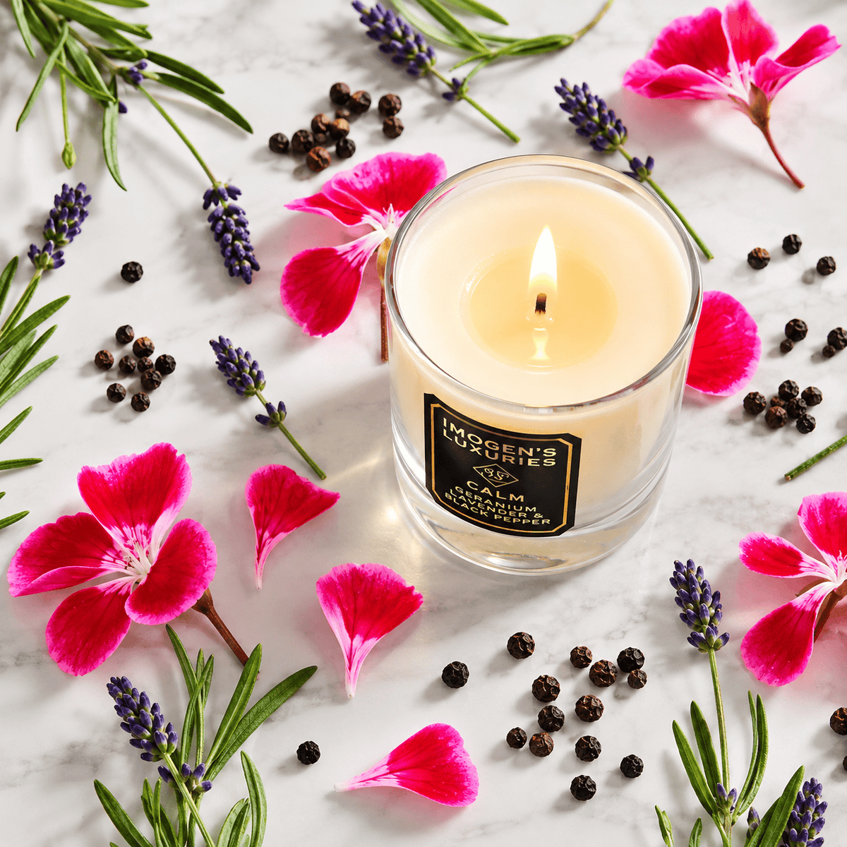 Image shows a top down angled image of a glass container candle with cream coloured wax and a lit flame with a pool of wax around the flame. The label says "IMOGEN'S LUXURIES CALM | Geranium, Lavender and Black Pepper Essential oils. Surrounding the candle are geranium leaves, sprigs of Lavender and black peppercorns.