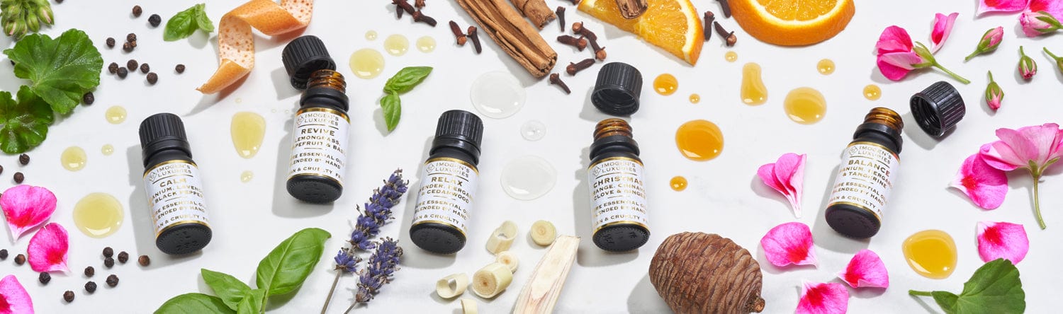 Image shows 5 black 10ml essential oil bottles with black caps. Bottles are surrounded with geranium petals, lemongrass strips, basil leaves and black peppercorns. These fruits and florals represent the ingredients in our essential oil blends.