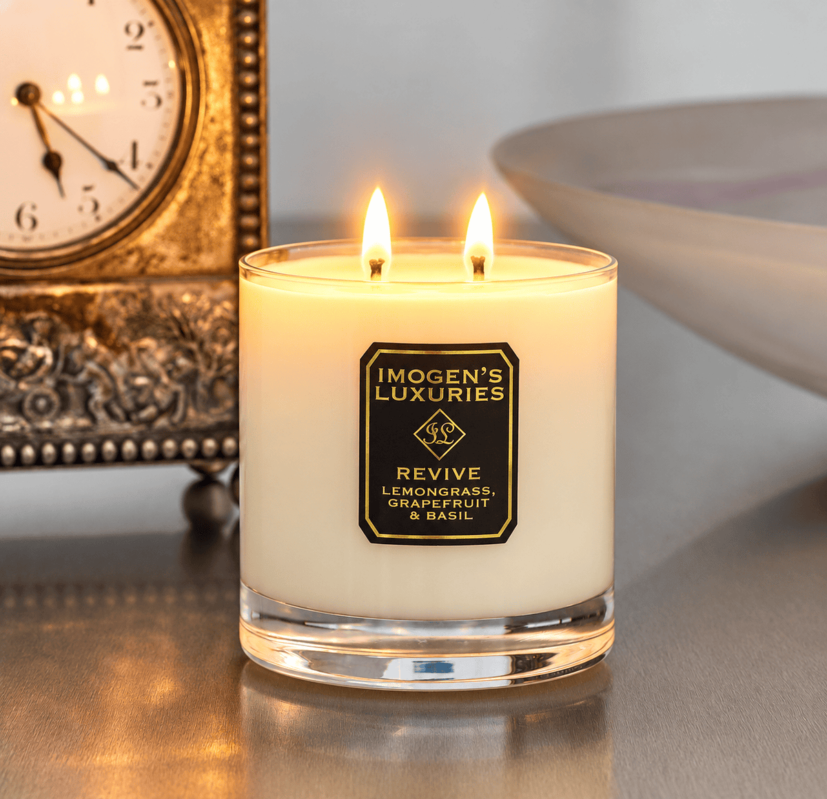 Cream coloured natural wax candle in glass container. Candles is scented with Lemongrass, Grapefruit & Basil Essential Oils. On the glass container is a black and gold label saying "Imogen's Luxuries" "Revive" The candle is burning with 2 cotton wicks showing atmospheric flames. The candle is sat in front of a carriage clock.