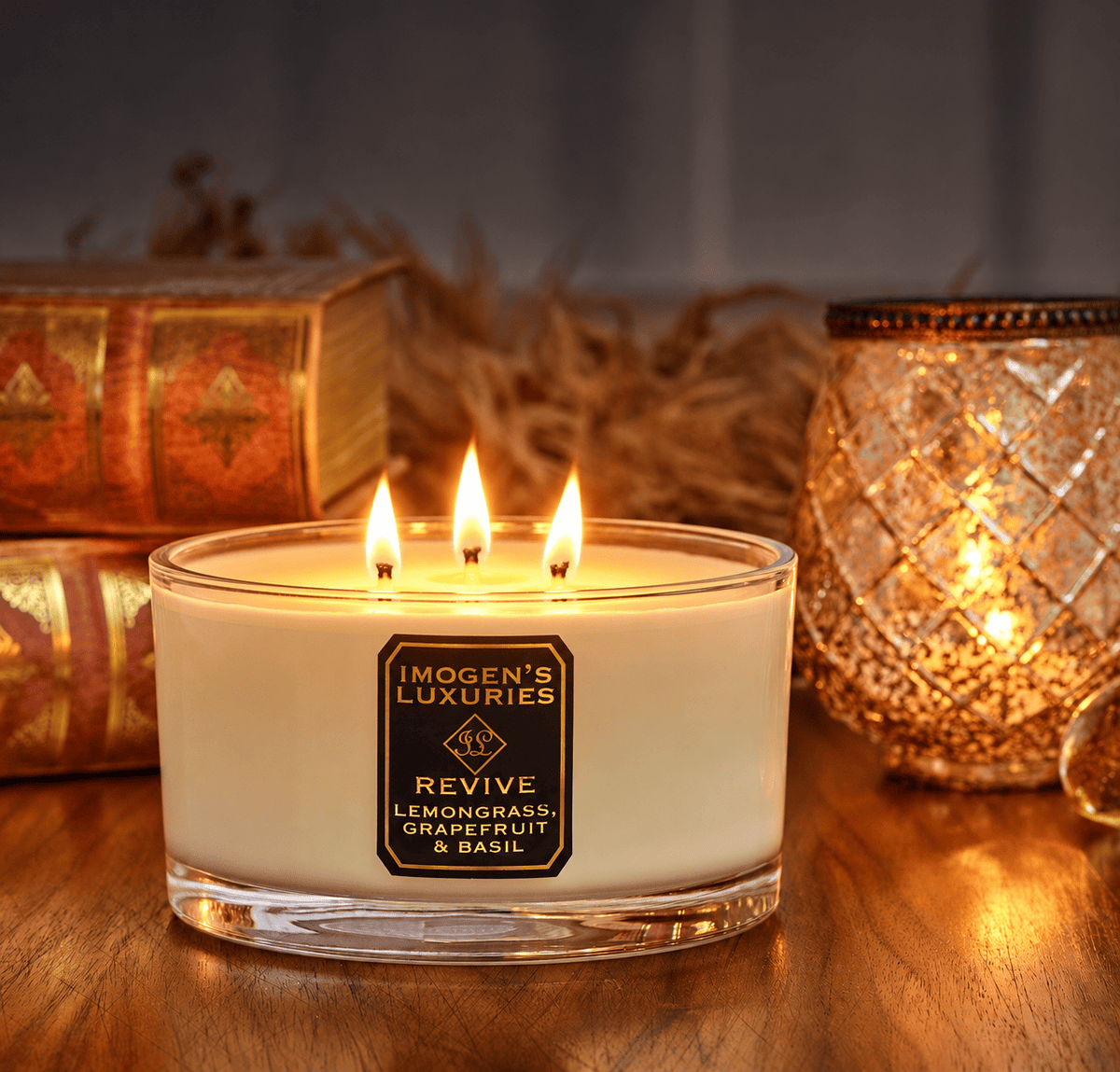 Imogen's Revive 3 wick candle burning in front of some books and an ornament, candle is set on a warm wood table.