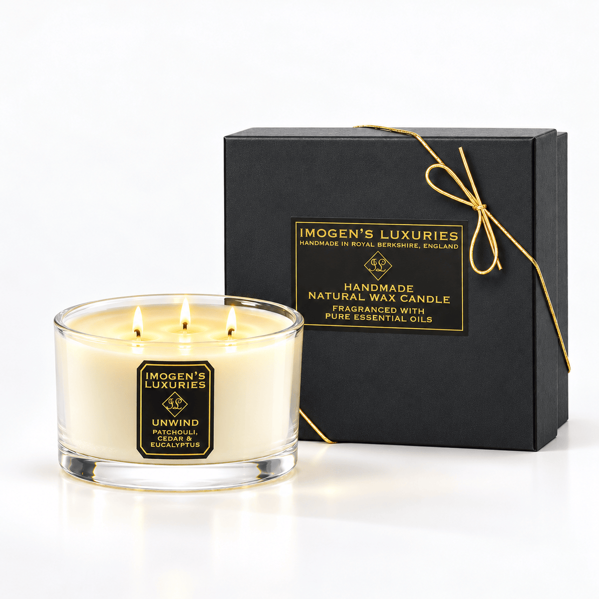 Cream coloured natural wax candle in glass container with 3 wicks which are ll lit. Candles is scented with Patchouli, Cedarwood & Eucalyptus Essential Oils. On the glass container is a black and gold label saying "Imogen's Luxuries" "Unwind". The candle sits by a solid black and gold box with a black and gold label saying "Handmade Natural Wax Candle fragranced with pure essential oils. There is a fine gold bow across the box.