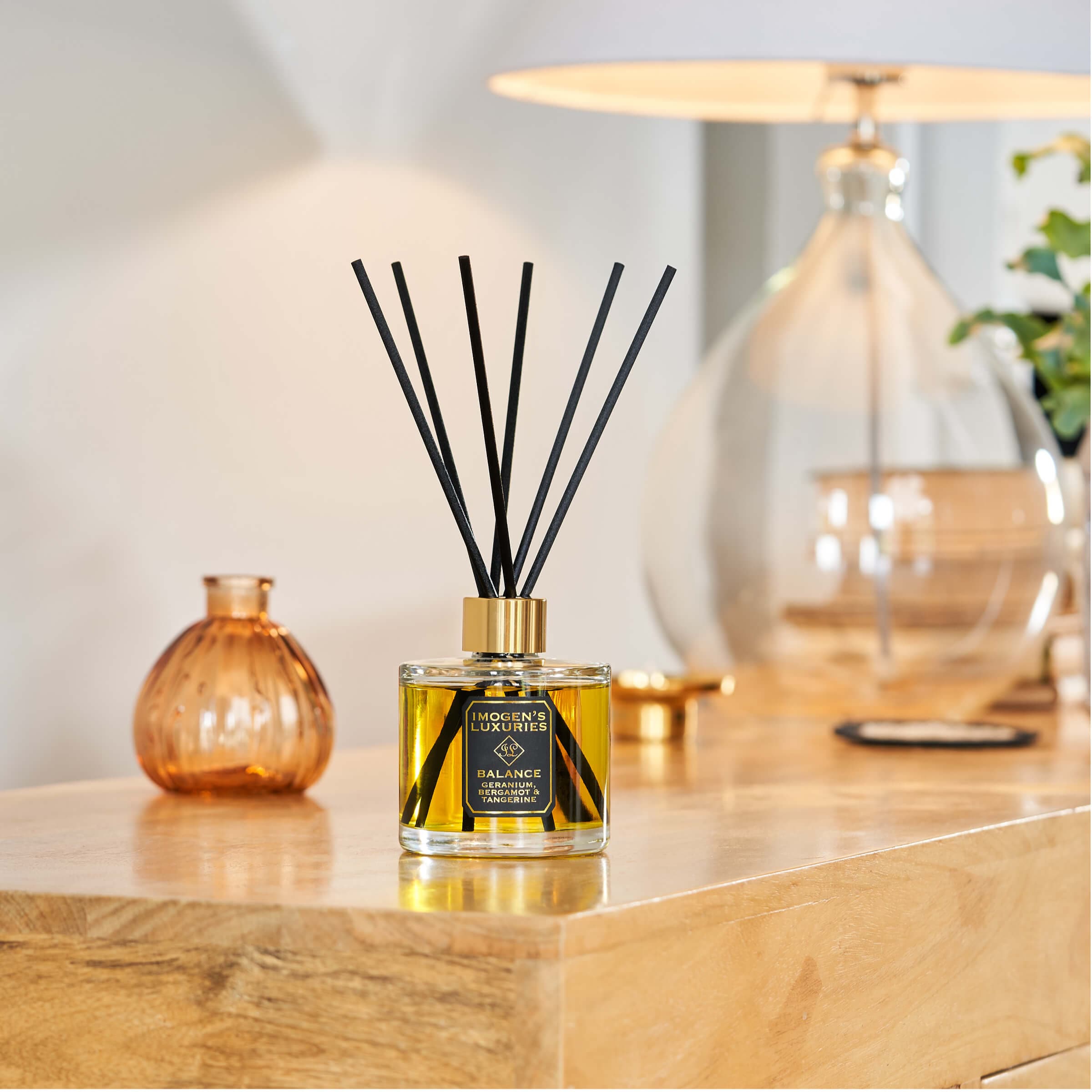 Balance luxury reed diffuser, large 200ml size with thick black reeds. Diffuser is placed on a wooden sideboard with a lamp slightly behind it. The scent is Geranium, Bergamot & Tangerine Essential Oils.