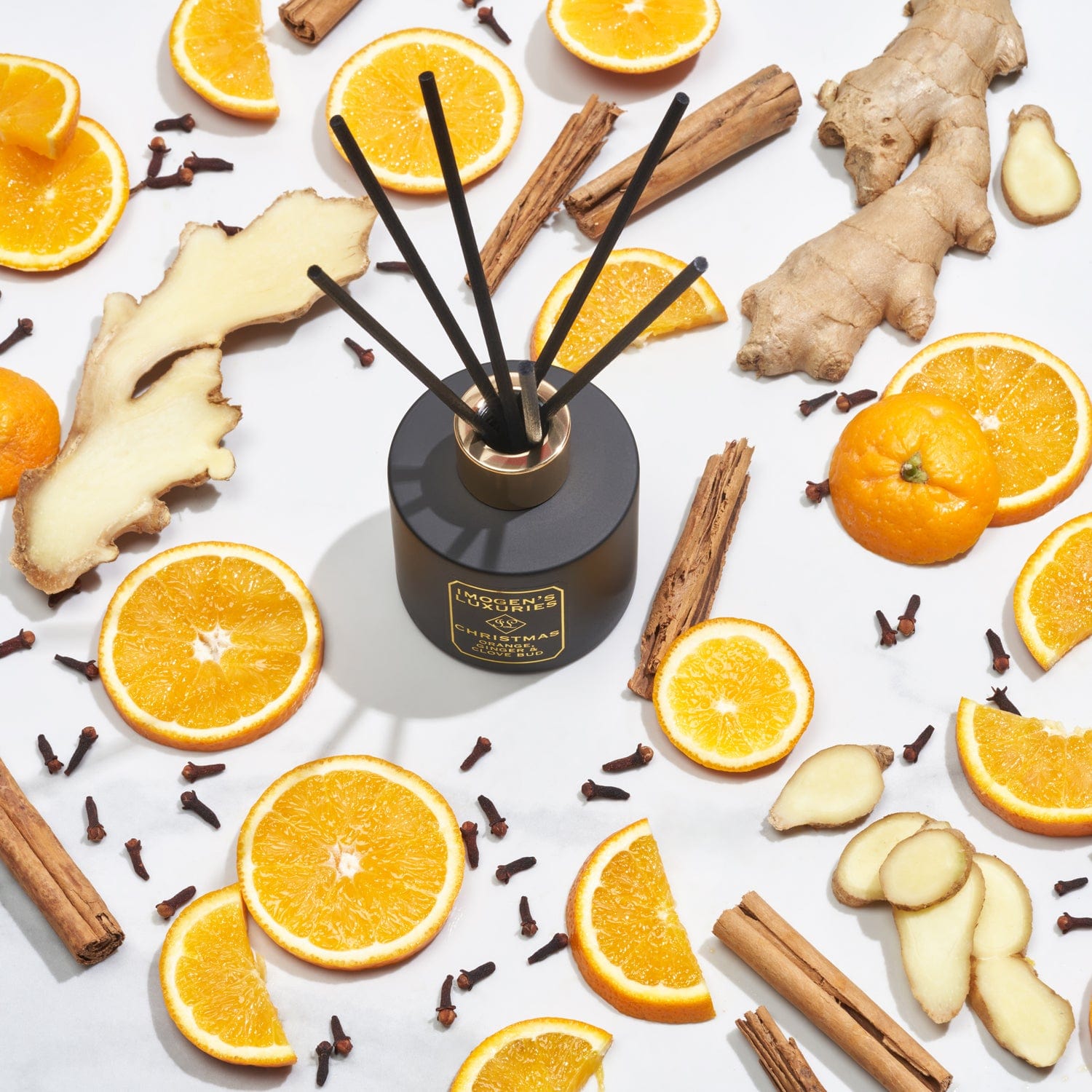 Christmas luxury black 100ml reed diffuser features a matte black 100ml bottle with 6 thick black reeds. The gold collar on the bottle and gold foil on the black label is elegant, luxurious and unisex.  Highly scented with Orange, Cinnamon, Clove and Ginger essential oils. Diffuser is shown surrounded by fruit and spices. Handmade by Imogen's Luxuries, Berkshire. UK. Natural, vegan and always cruelty free.