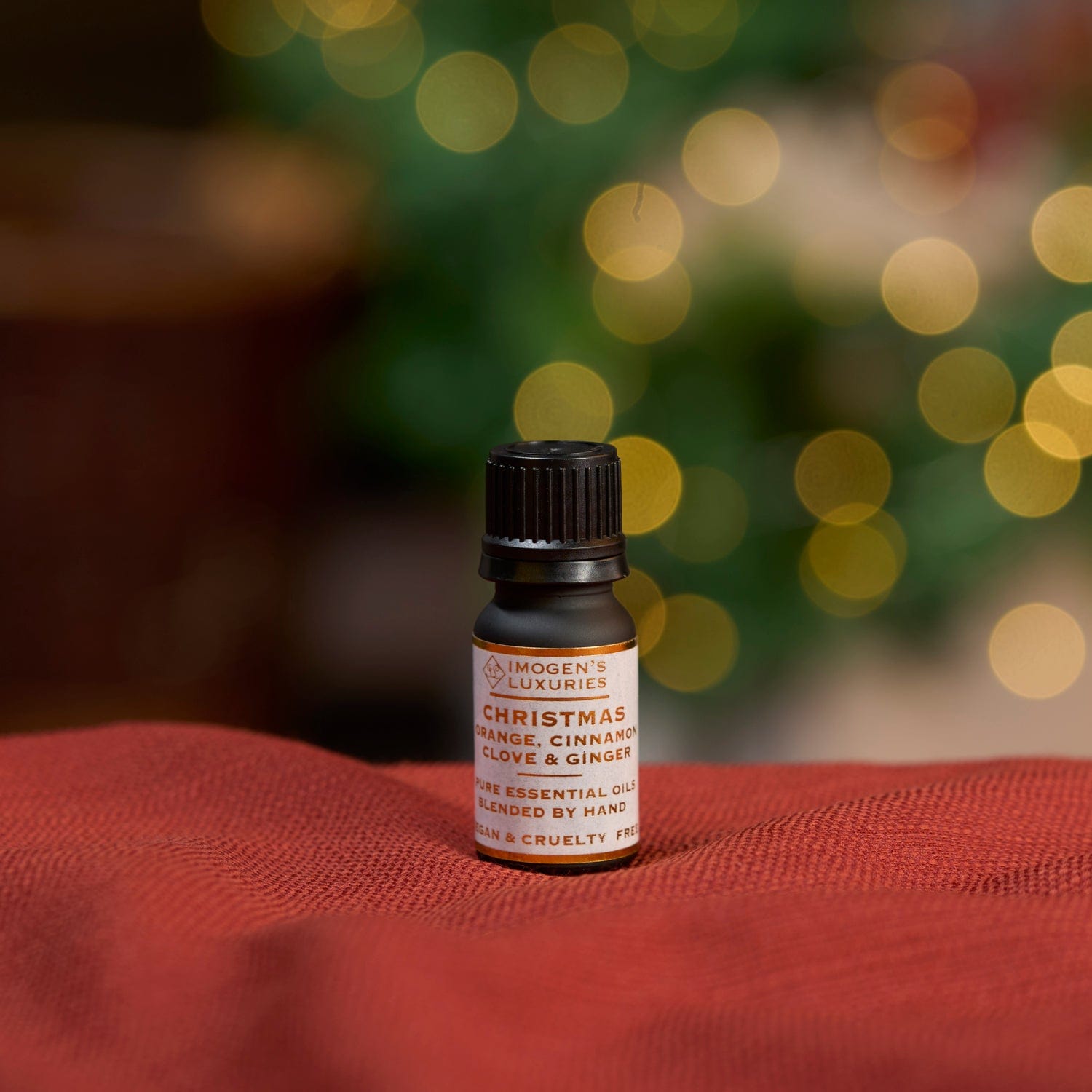 Award-winning Christmas blend of pure essential oils. Highly concentrated Orange, Cinnamon, Clove and Ginger oils in a 10ml black dropper bottle with white and gold label. Bottle is shown resting on a red cloth with Christmas lights in background. Imogen's Luxuries, Berkshire