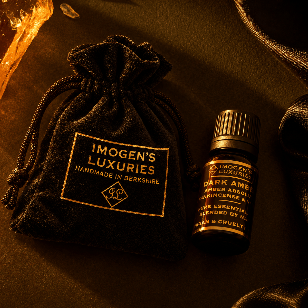 A 10ml blck glass bottle of Imogen's Luxuries Dark Amber  with a black cap, displayed next to a matching black velvet drawstring pouch with gold lettering, on a black textured background. The label says "IMOGEN'S LUXURIES DARK AMBER, AMBER ABSOLUTE, FRANKINCENSE AND VETIVER"