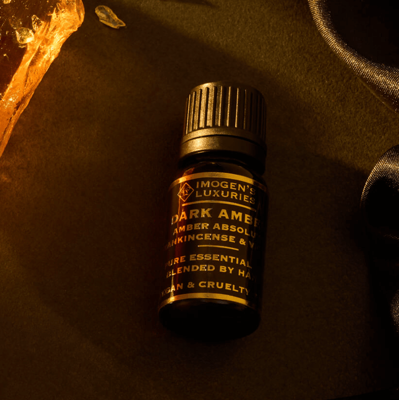 Imogen's Luxuries 10ml black dropper bottle containing Dark Amber Arabian Oud Essential oil blend. Notes of Amber Absolute, Vetiver and Frankincense beside a large piece of Amber resin on a dark silk surface. All Natural Essential Oils. Vegan and Cruelty Free.