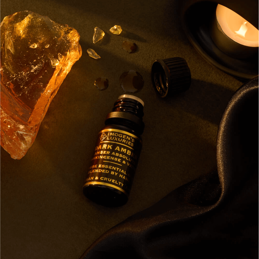 10ml Black dropper bottle of Imogen's Luxuries Dark Amber Oud Oil Blend. Notes of Amber Absolute, Vetiver and Frankinsence. Bottle is beside Amber resin on a dark background with an oil burner.