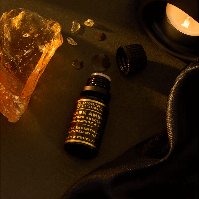 10ml Black dropper bottle of Imogen's Luxuries Dark Amber Oud Oil Blend. Notes of Amber Absolute, Vetiver and Frankinsence. Bottle is beside Amber resin on a dark background with an oil burner.