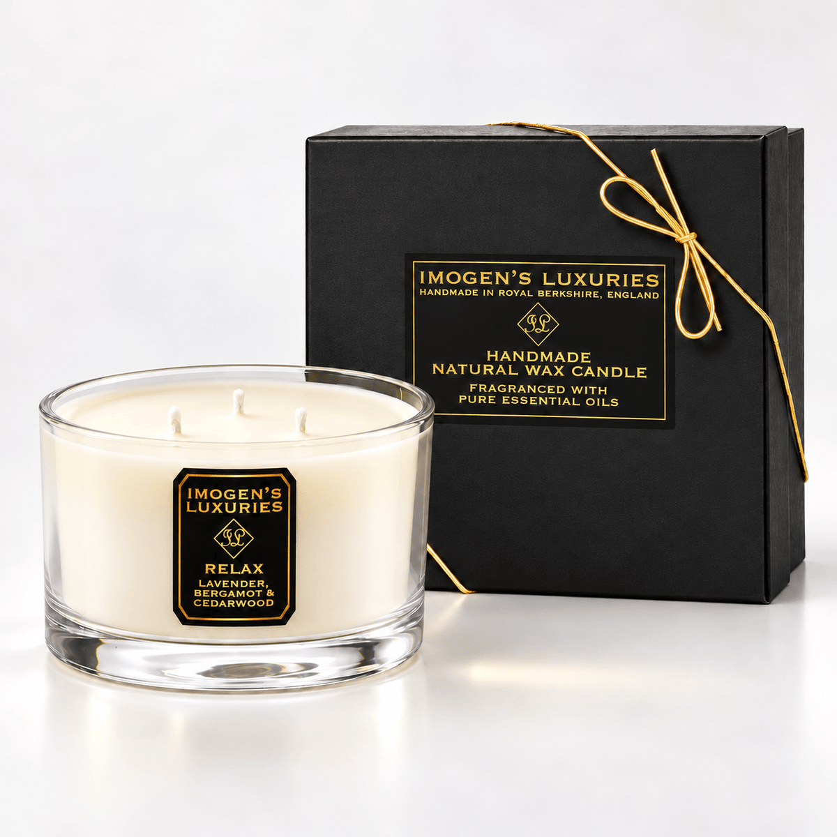 Cream coloured natural wax candle in glass container with 3 wicks which are all unlit. Candles is scented with Lavender, Bergamot & Cedarwood Essential Oils. On the glass container is a black and gold label saying "Imogen's Luxuries" "RELAX". The candle sits by a solid black and gold box with a black and gold label saying "Handmade Natural Wax Candle fragranced with pure essential oils. There is a fine gold bow across the box.