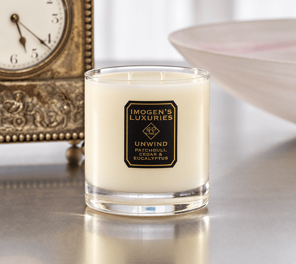 Cream coloured natural wax candle in glass container. Candles is scented with  Patchouli, Cedarwood & Eucalyptus  Essential Oils. On the glass container is a black and gold label saying "Imogen's Luxuries" "Unwind". The candle is sat in front of a carriage clock.