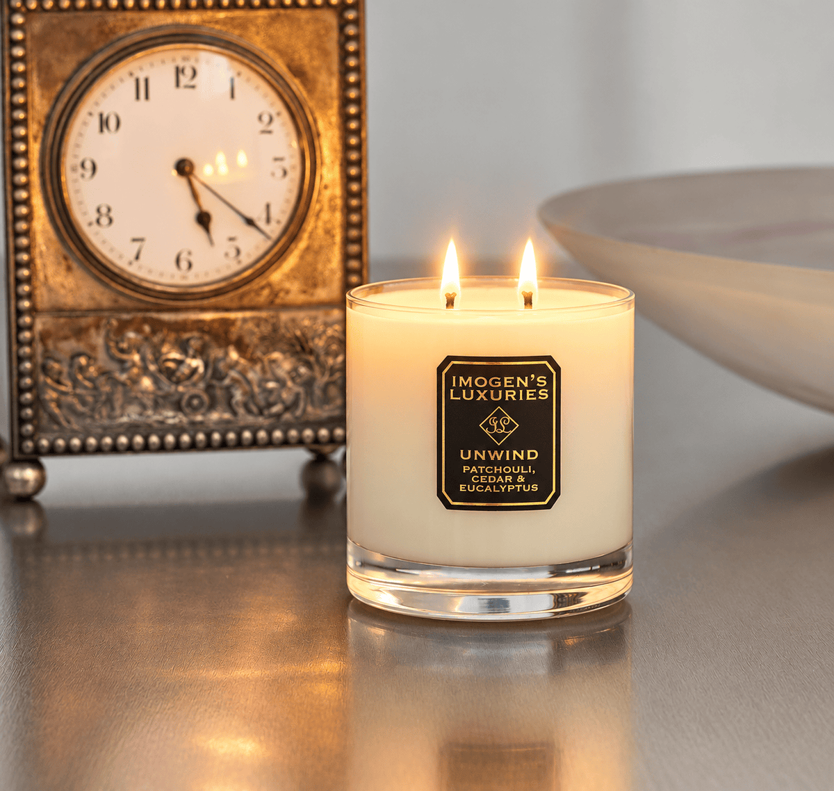Cream coloured natural wax candle in glass container. Candles is scented with  Patchouli, Cedarwood & Eucalyptus  Essential Oils. On the glass container is a black and gold label saying "Imogen's Luxuries" "Unwind" The candle is burning with 2 cotton wicks showing atmospheric flames. The candle is sat in front of a carriage clock.