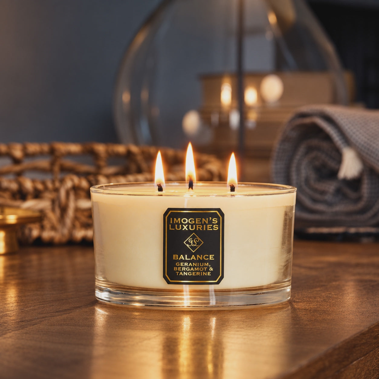 Video of a flickering 3 wick candle in a glass container with a black and gold label which reads "IMOGEN'S LUXURIES BALANCE, GERANIUM, BERGAMOT & TANGERINE. Candle has all 3 wicks lit and is sat on a sideboard made of dark wood with a lamp and a grey throw in the background 
